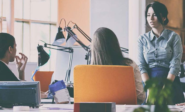 dark haired woman sitting on desk talking to coworkers